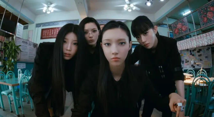  ILLIT members with long black hair, dressed in black outfits, looking intently at the camera in a colorful restaurant setting with teal chairs and vibrant decor.