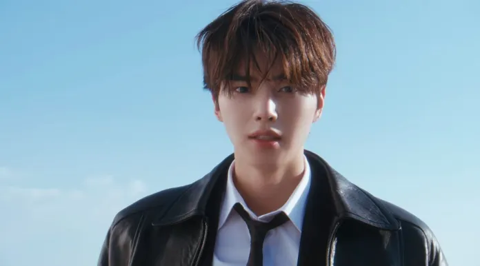 Song Kang with tousled hair wearing a black leather jacket and a white shirt stands outdoors against a clear blue sky.