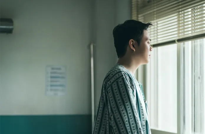 A young man in a striped outfit standing by a window, looking out thoughtfully.