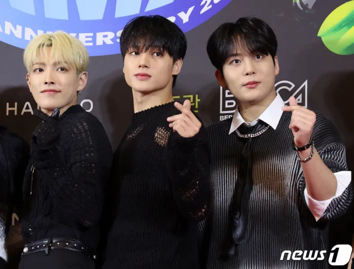 ATEEZ posing together on a red carpet, dressed in stylish black knitted sweaters. They are making gestures with their hands, smiling at the camera.