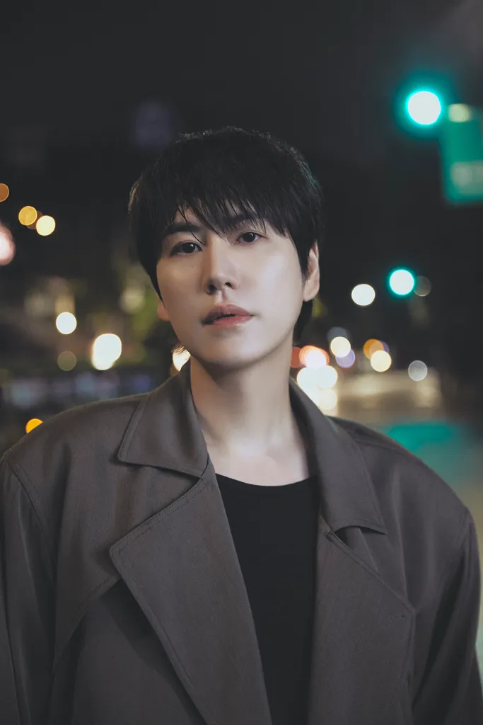 Kyuhyun with dark hair wearing a large coat, posing outdoors at night with blurred city lights in the background.