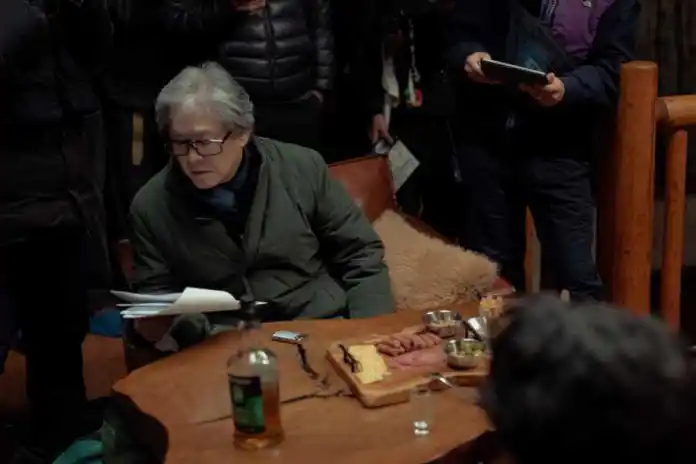 A group of people gathered around a wooden table, with an older man reading scripts while sitting on a chair. The table features various food items and drinks, and others in the background observe and take notes.