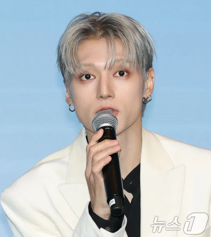 ATEEZ with silver hair wearing a white blazer, holding a microphone and speaking during a press event.