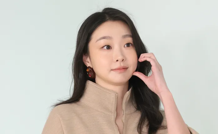 Kim Da-mi with long black hair, wearing a tan high-collared top and colorful earrings, smiling and touching her face against a light background.