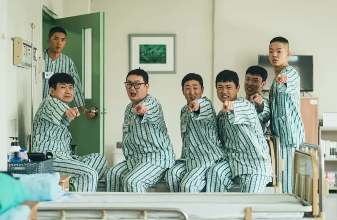 A group of seven men wearing matching hospital pajamas are playfully pointing at the camera, with expressions of surprise and excitement. The scene takes place in a hospital room, featuring a bed and medical equipment.