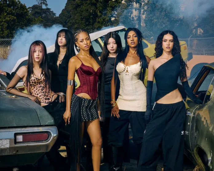 KATSEYE posing together in front of vintage cars, with varying fashion styles, set against a backdrop of smoke and greenery.
