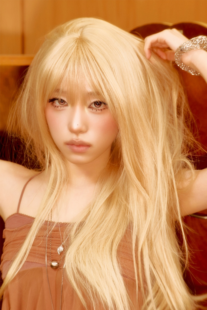 YENA with long, blonde hair styled casually, wearing a brown top, posing in a relaxed setting.
