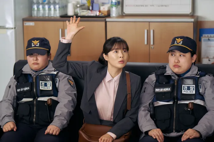 A woman in a business suit raises her hand while sitting between two police officers, who are wearing uniforms and sitting on a couch in a police station.