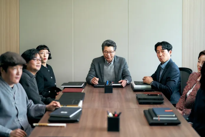 A group of six people sitting around a conference table in a meeting room, with documents and stationery on the table. A man in the center looks engaged while others display various expressions.