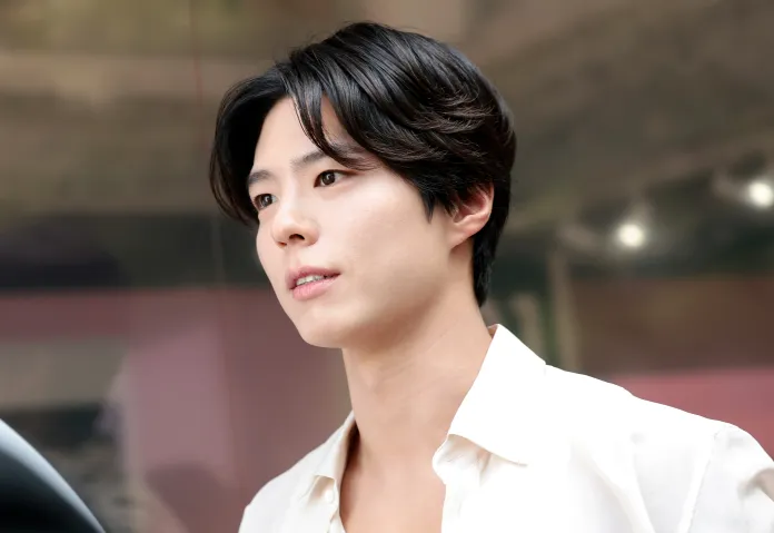 A young man with dark, wavy hair wearing a white shirt, looking slightly away with a thoughtful expression against a blurred background.