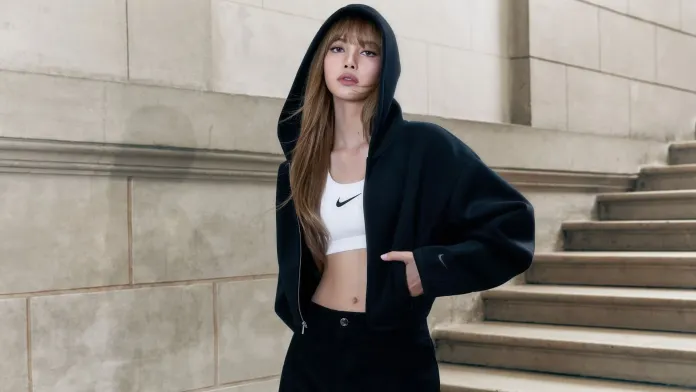 A fashionable woman wearing a black hoodie over a white crop top, standing on a staircase with a neutral background.