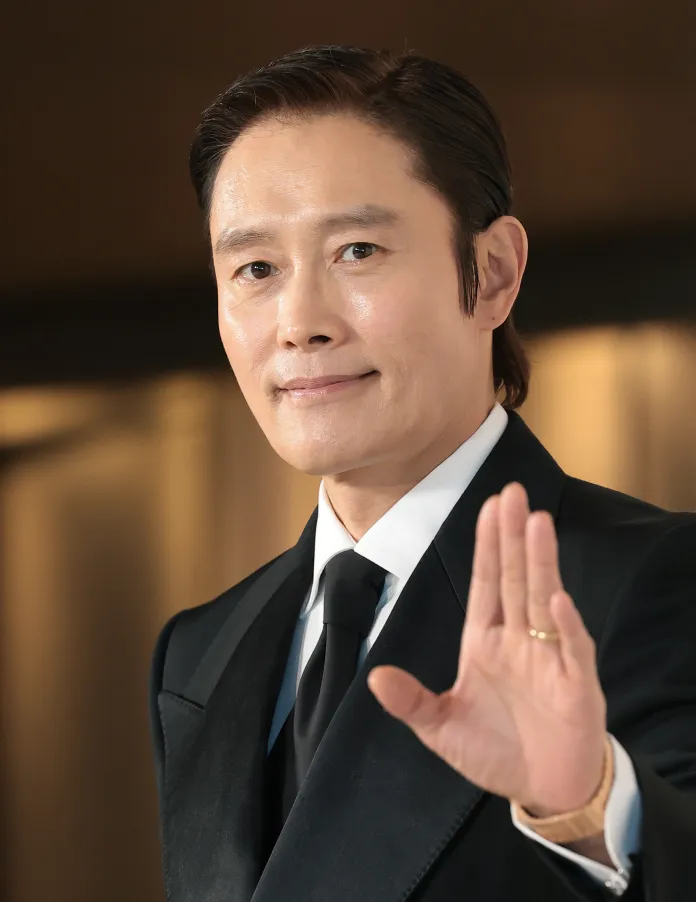 Lee Byung-hun  in a formal black suit giving a friendly gesture with his right hand raised, smiling at the camera.