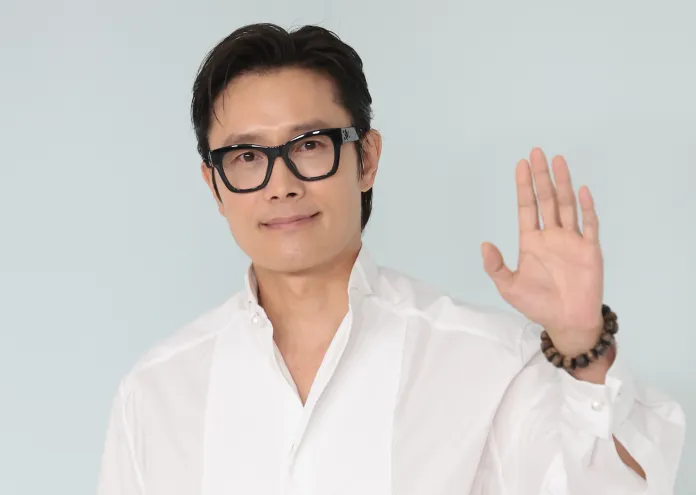 Lee Byung-hun  in a white shirt with a high collar and black glasses is smiling and waving at the camera, set against a light blue background.