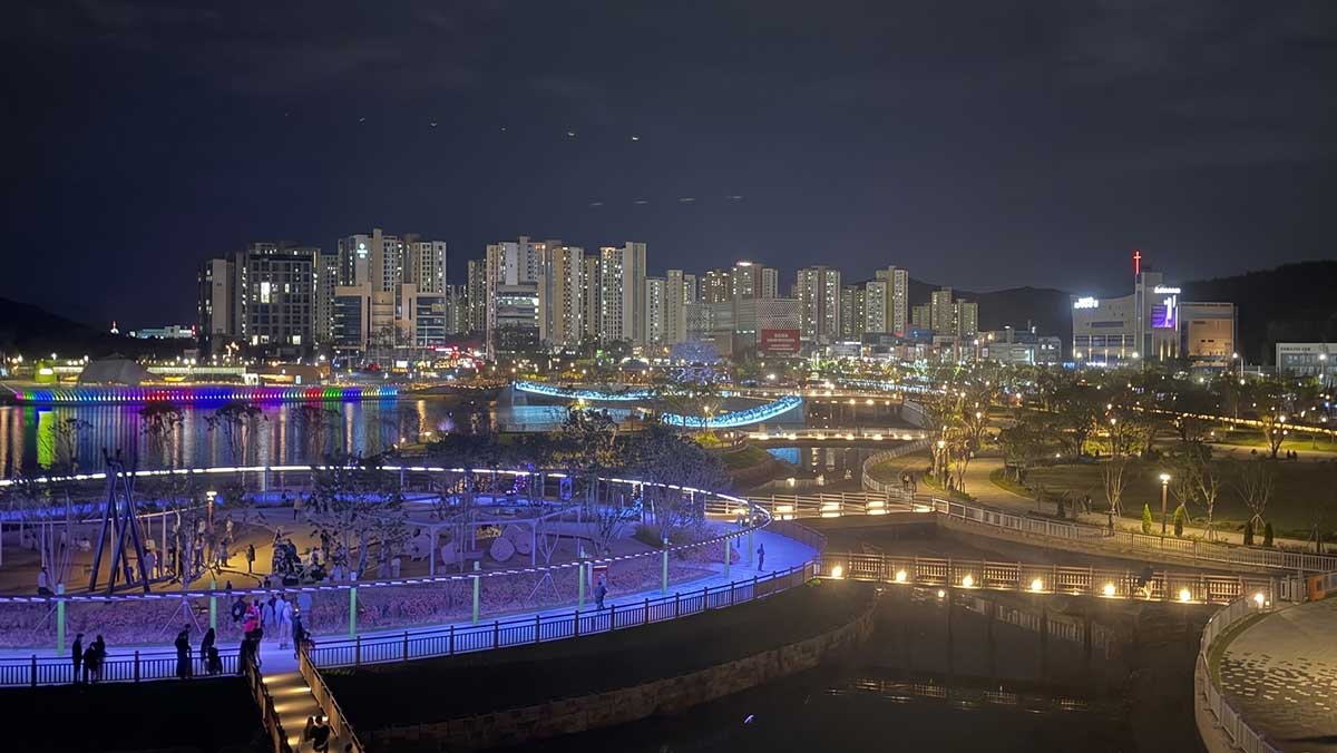 호수 주변 산책로와 출렁다리, 놀이 공간에 조명이 켜지고 도안신도시 아파트 단지가 뒤로 보인다. / 대전 갑천 생태호수공원, ai