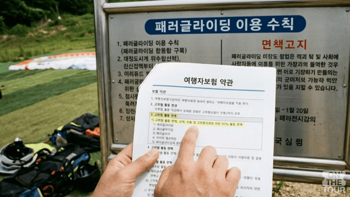 패러글라이딩 이용 수칙 안내판 앞에서 여행자보험 약관의 '고위험 활동 면책' 조항을 손가락으로 가리키고 있다. (AI 생성) / 온더투어