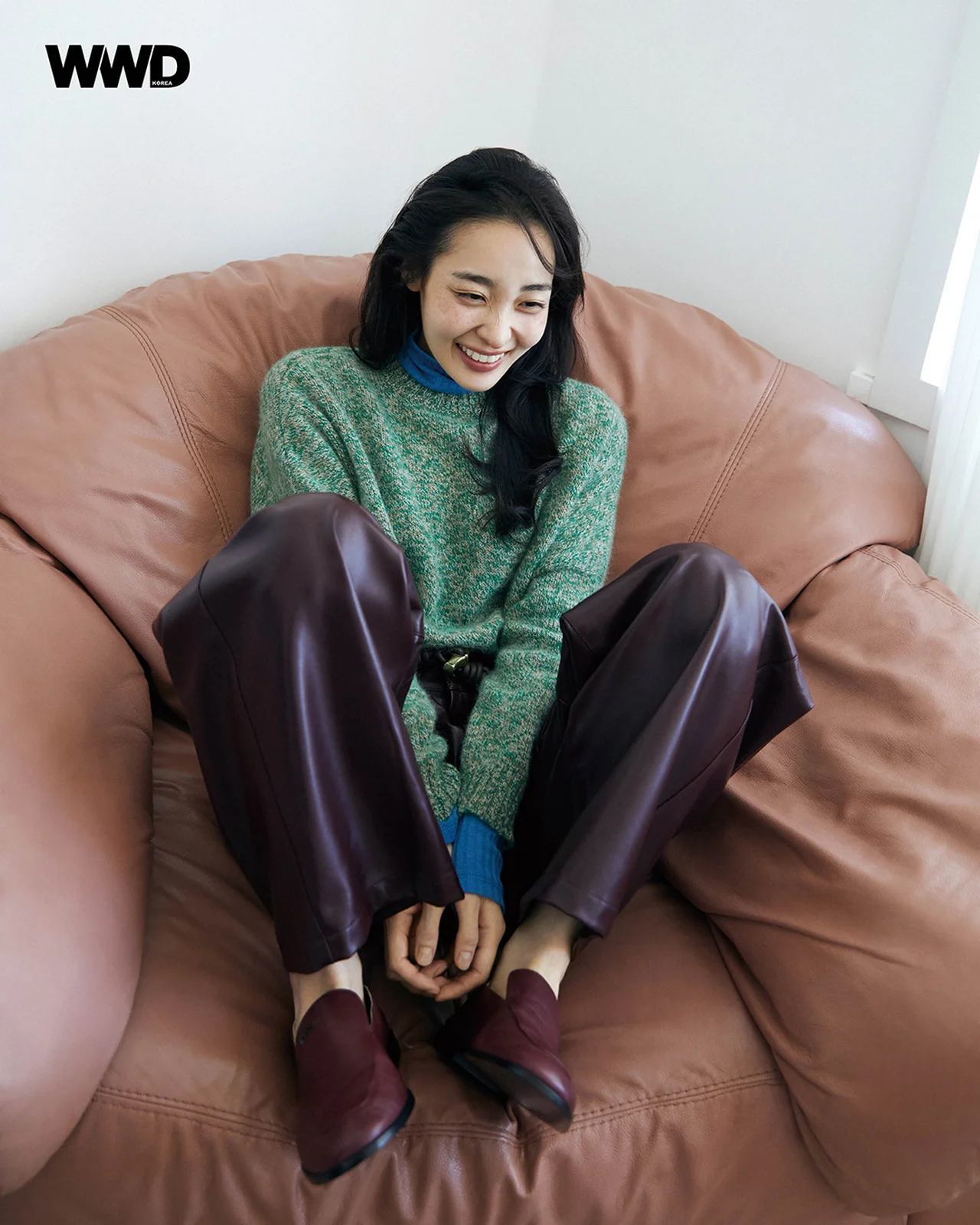 연두색 니트와 버건디색 가죽 팬츠를 입고 갈색 소파에 앉아 환하게 웃고 있는 김민하. / 사진=WWD KOREA
