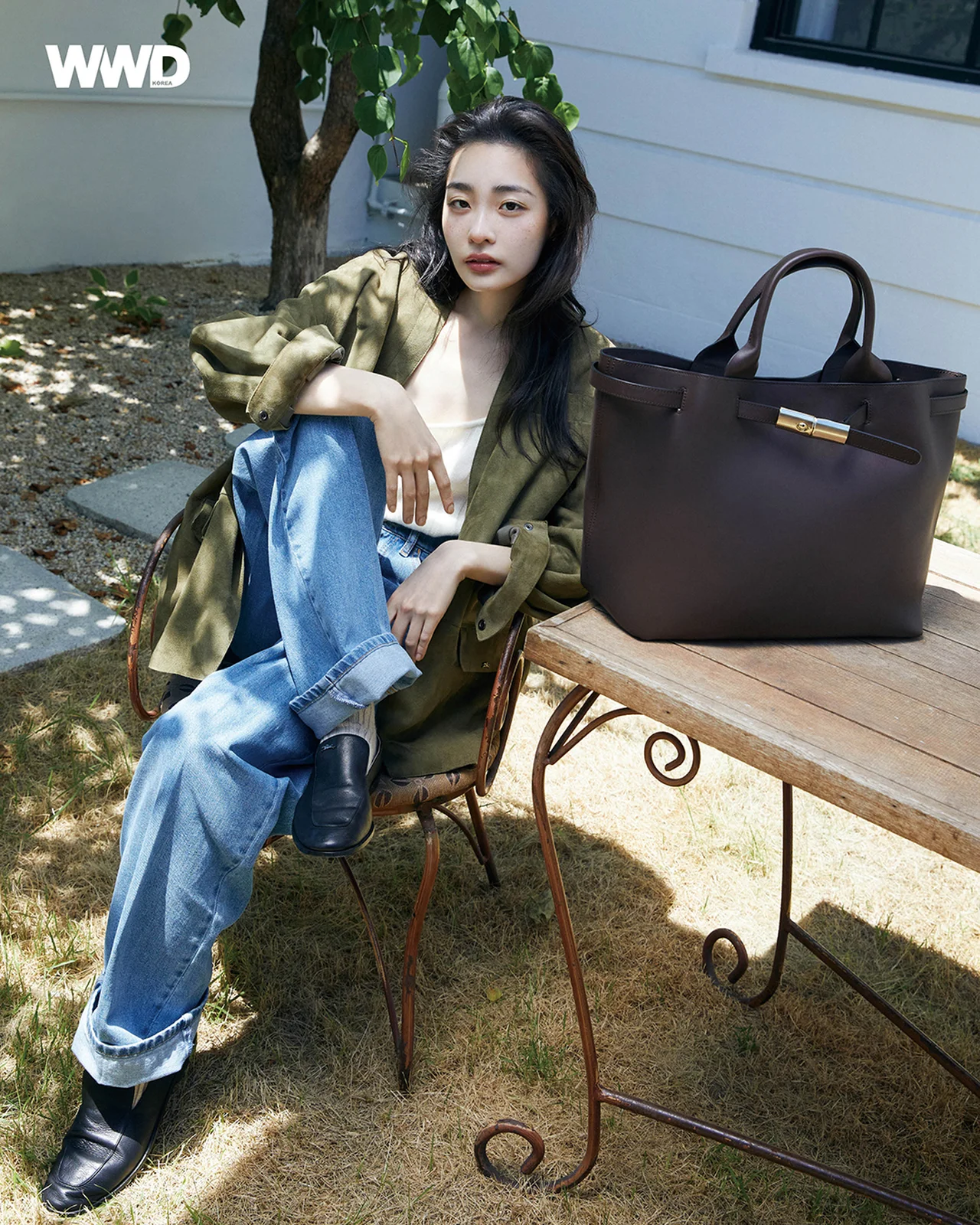 카키색 재킷과 롤업 청바지를 입고 야외 테이블에 앉아 있는 김민하가 어두운 갈색 토트백과 함께 포즈를 취하고 있다. / 사진=WWD KOREA