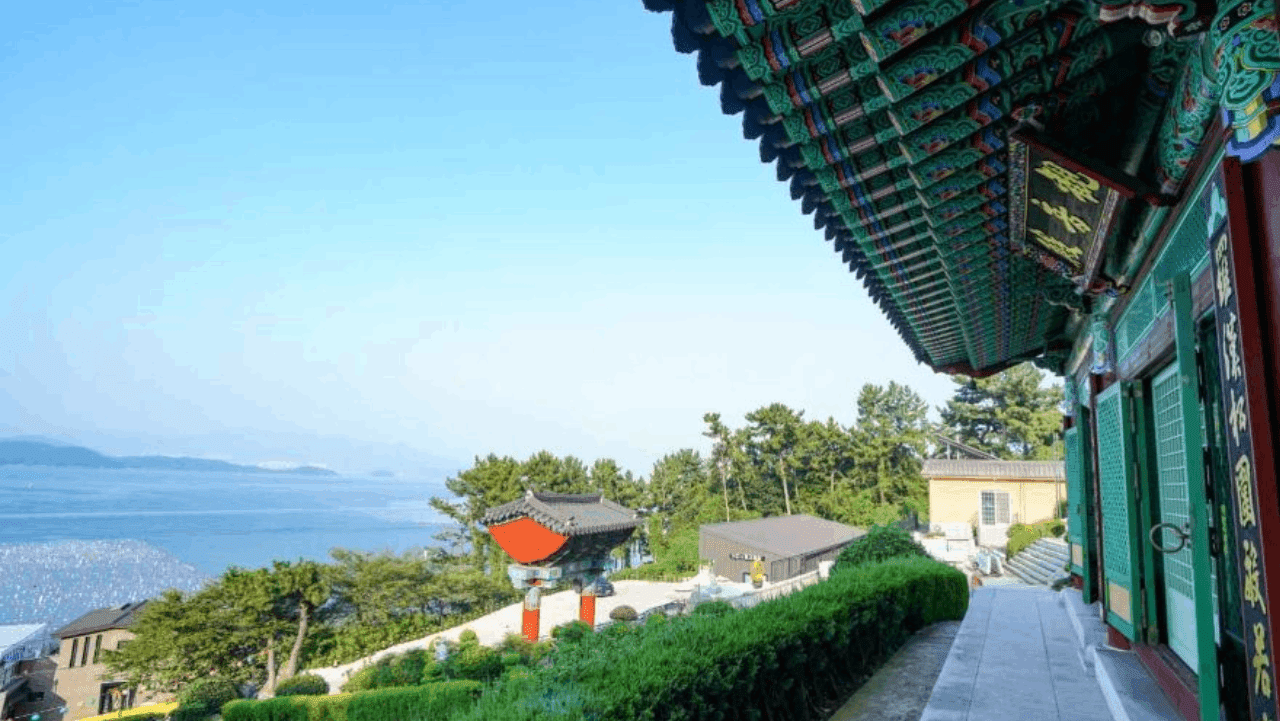 계단 끝 정상 전각 처마 아래에서 한눈에 내려다보는 정겨운 사찰 경내와 남해 수평선 풍경. / 한국관광공사