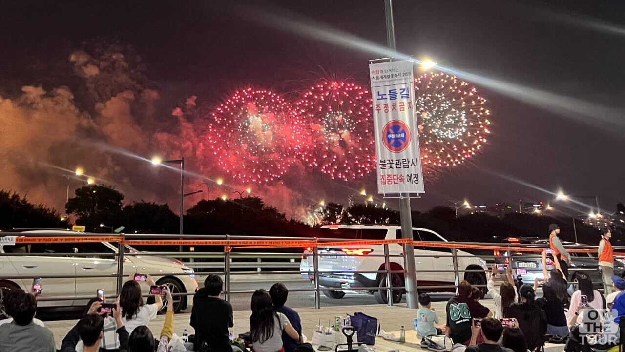 안전 펜스 뒤로 관람객들이 질서정연하게 자리를 잡고 붉은 불꽃 연출을 감상하는 현장 풍경. / 온더투어