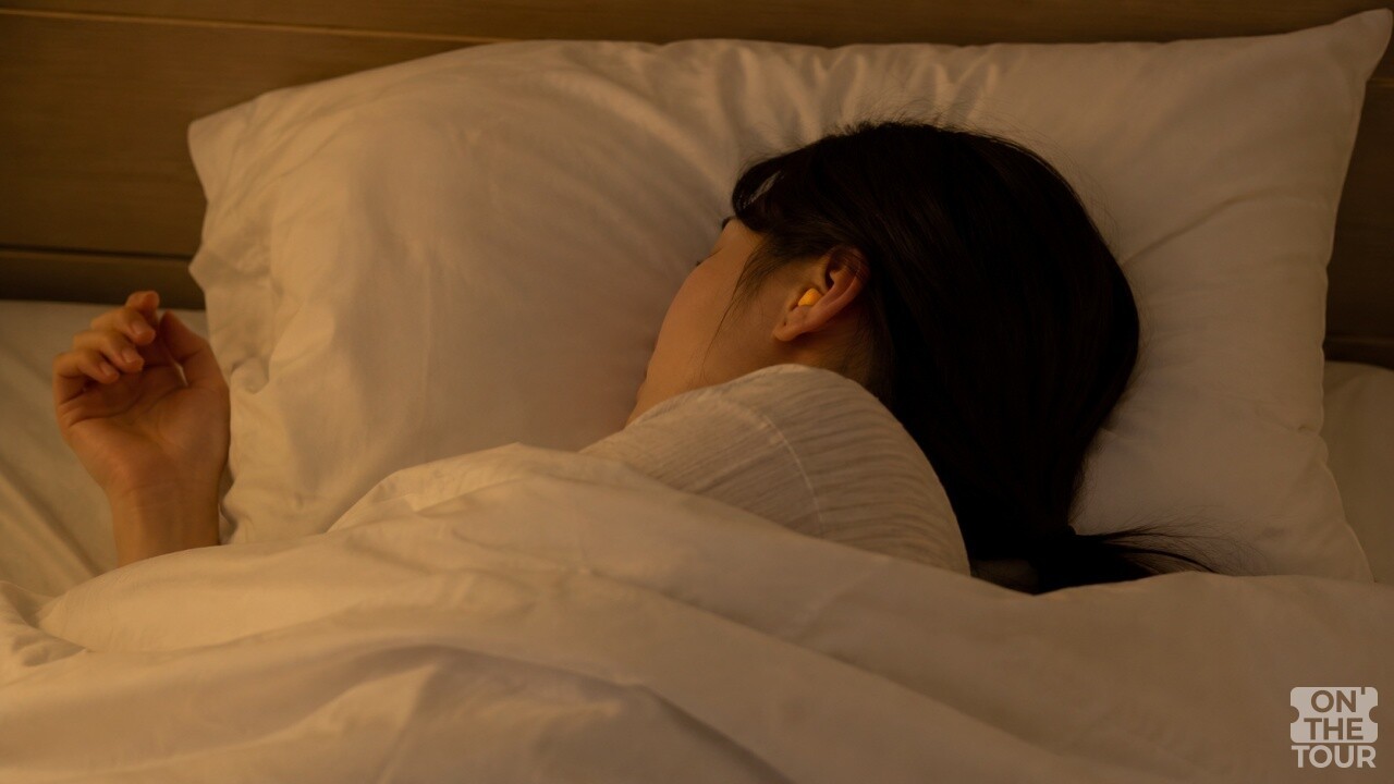 귀마개를 끼고 침대에 누워 있는 여성. (AI 생성) / 온더투어