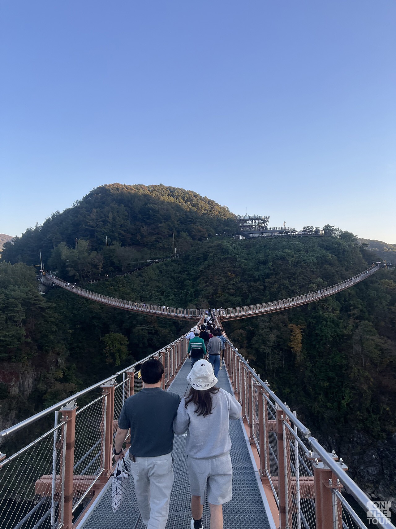 Y자로 갈라지는 다리 중앙을 향해 방문객이 걷고 있고, 뒤쪽 언덕 위에 전망대가 보인다. / 온더투어