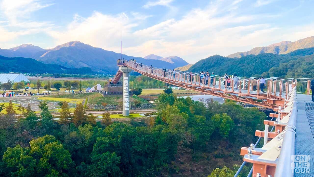 한탄강 출렁다리 위로 방문객이 걸어가고, 멀리 산자락과 들판이 이어진다. / 온더투어