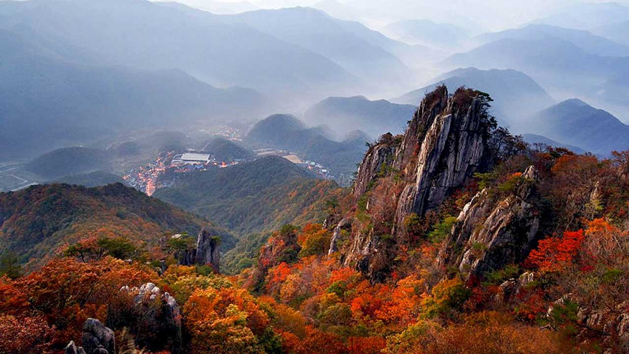 완주 대둔산 암봉 아래로 붉게 물든 단풍이 번져 있고, 멀리 산자락 사이로 마을이 내려다보인다. / 투어전북
