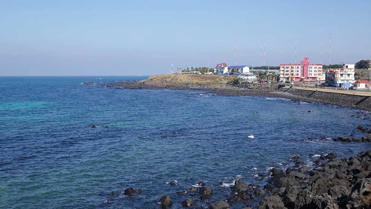 제주 애월해안도로 변 현무암 바위 해안과 푸른 바다, 멀리 보이는 해안 마을. / 비짓제주